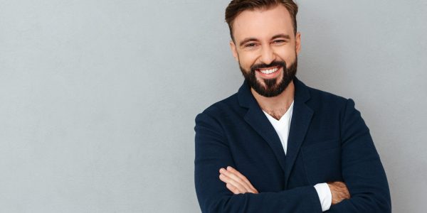 Happy bearded man in business clothes with crossed arms looking at the camera over gray background
