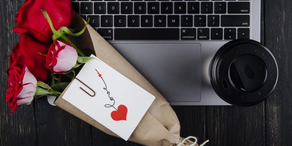 top view a bouquet of red roses in craft paper with attached postcard lying on a laptop with a paper cup of coffee on dark wooden background